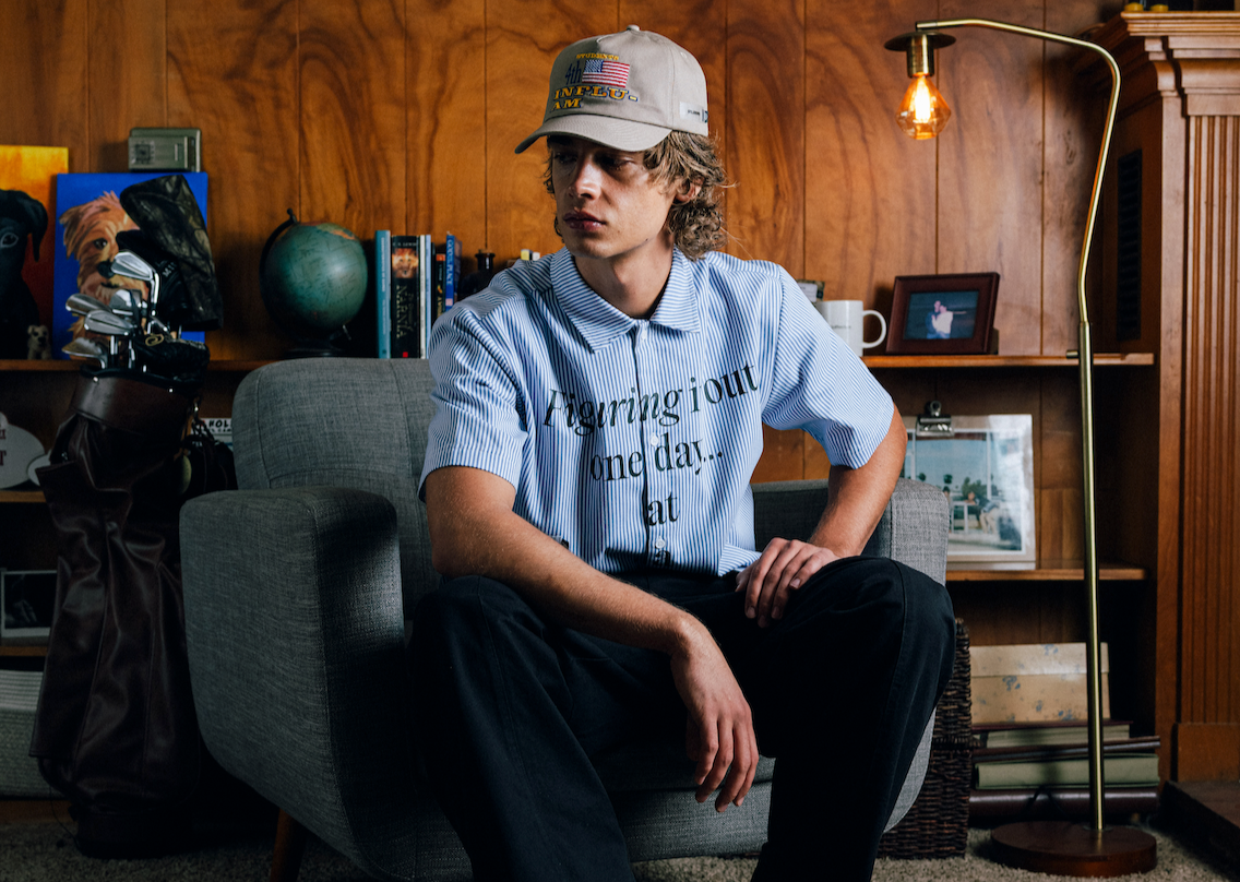 Person sitting on a couch wearing a cap and a shirt with text, in a room with wooden walls and shelves.
