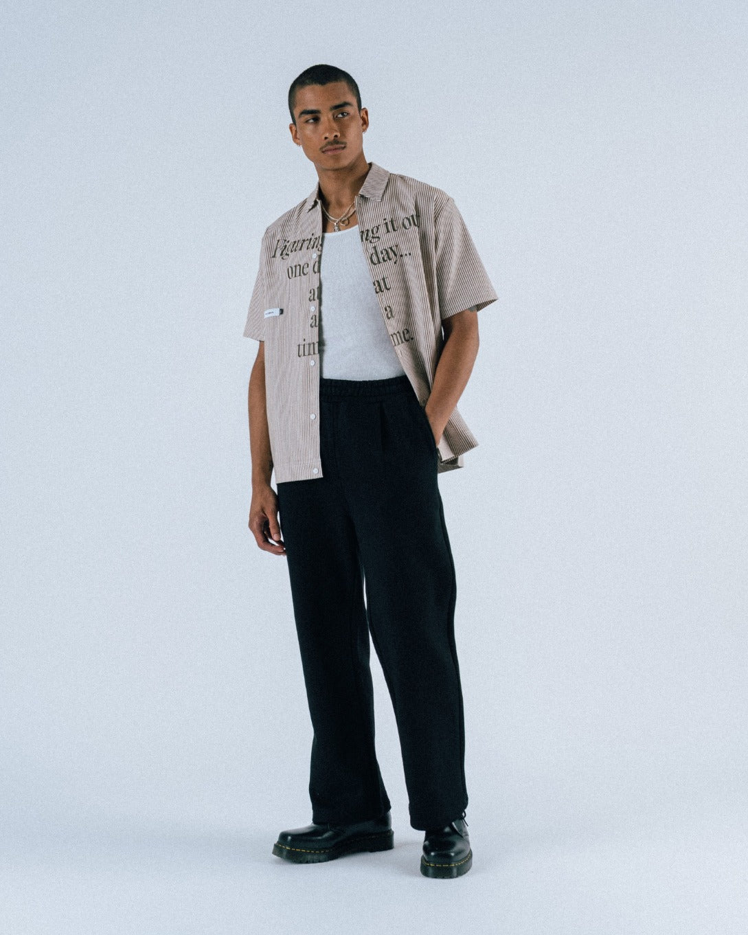 Man wearing a beige shirt with text, white tank top, black pants, and black shoes on a light gray background