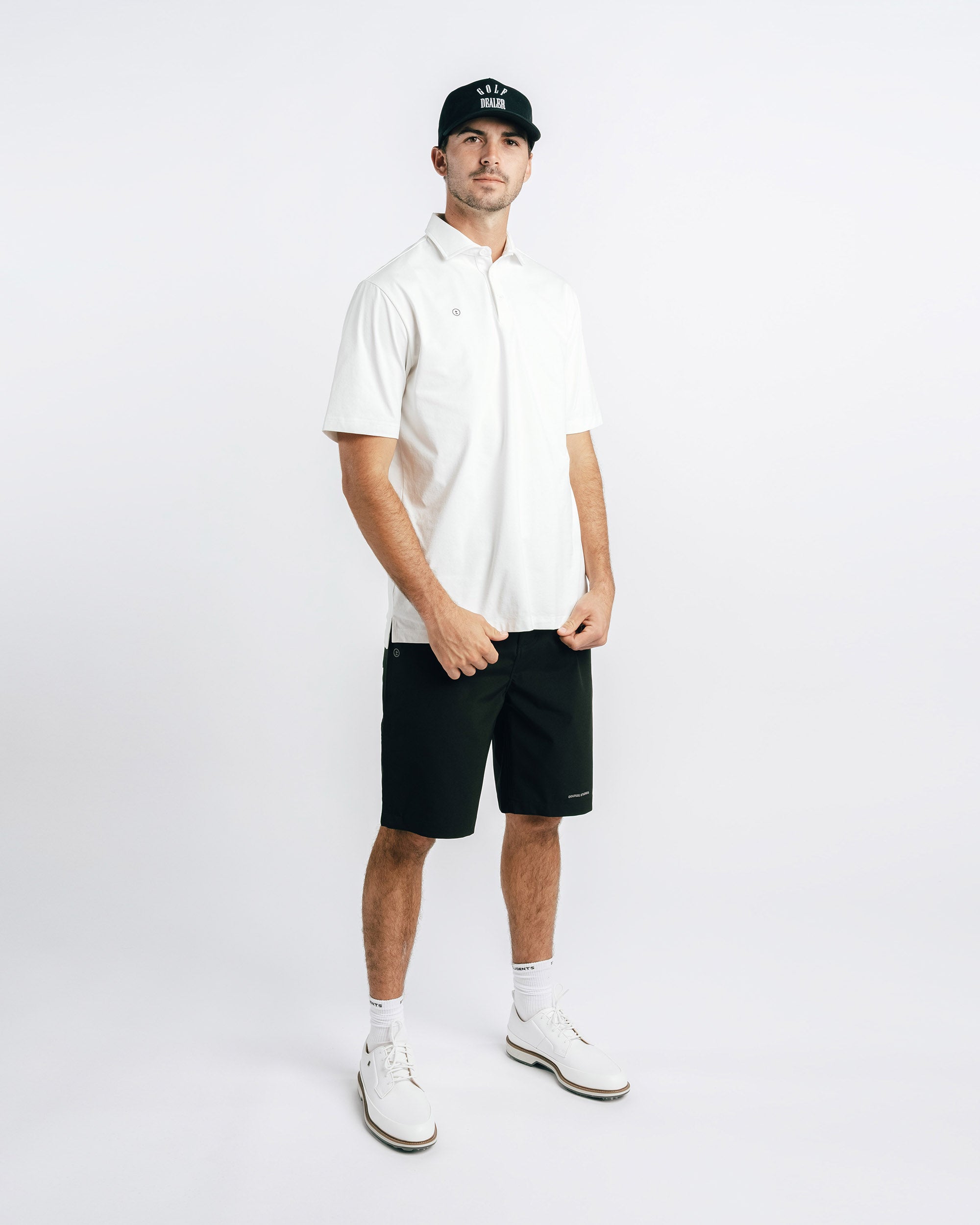 Man wearing a white polo shirt, black shorts, and a black cap on a white background
