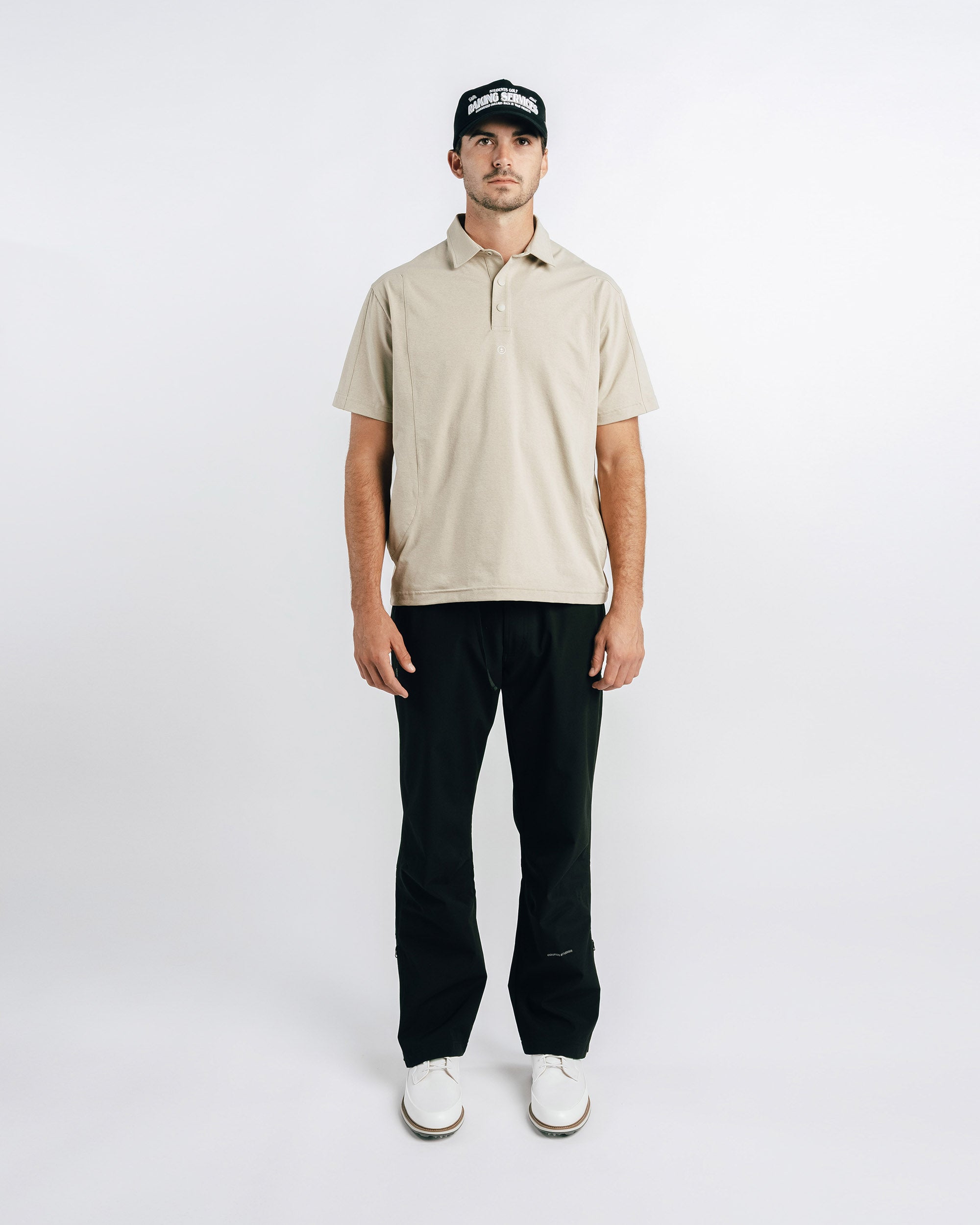 Man wearing a beige polo shirt, black pants, and a black cap on a white background