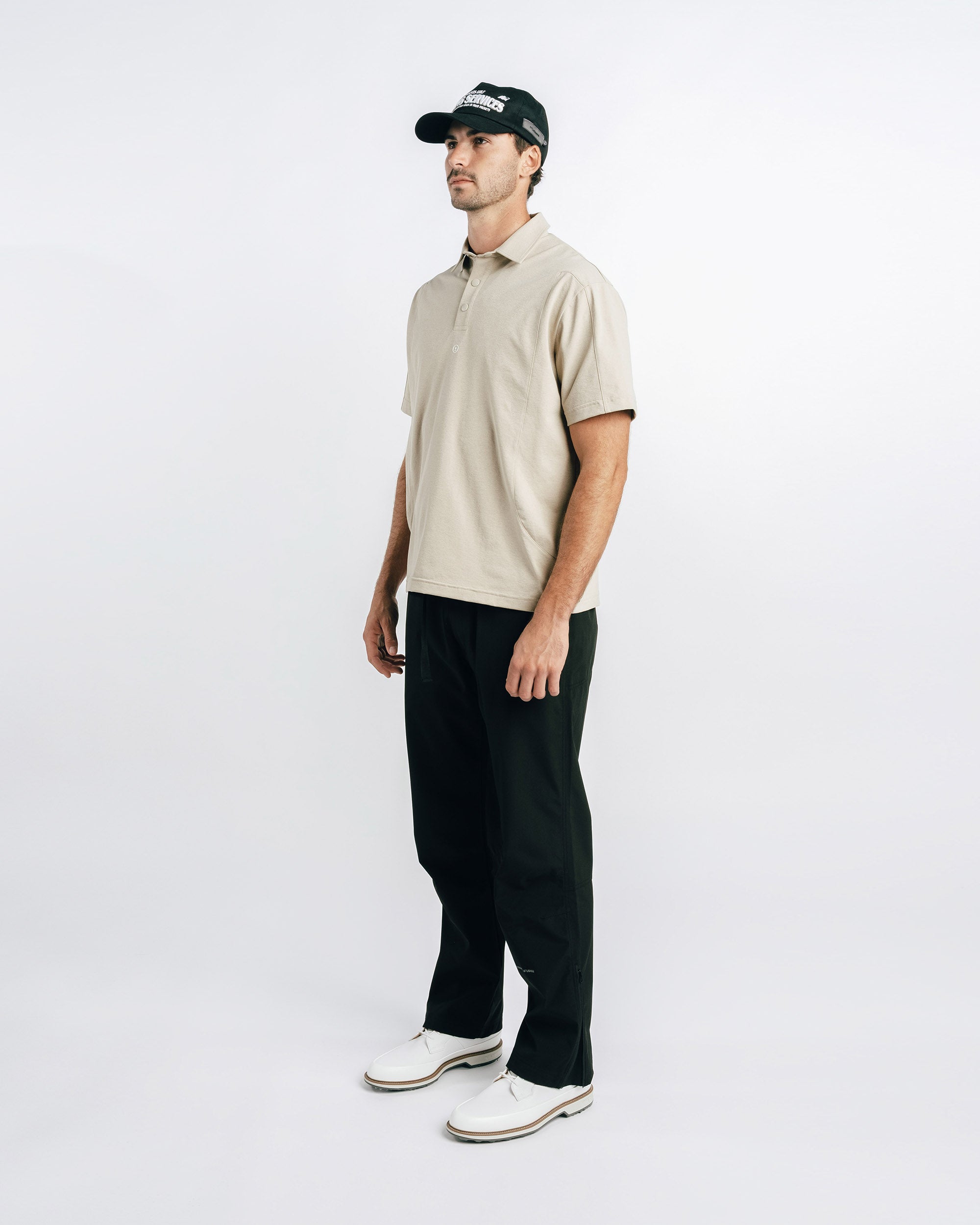 Man wearing a beige polo shirt, black pants, and a cap on a white background