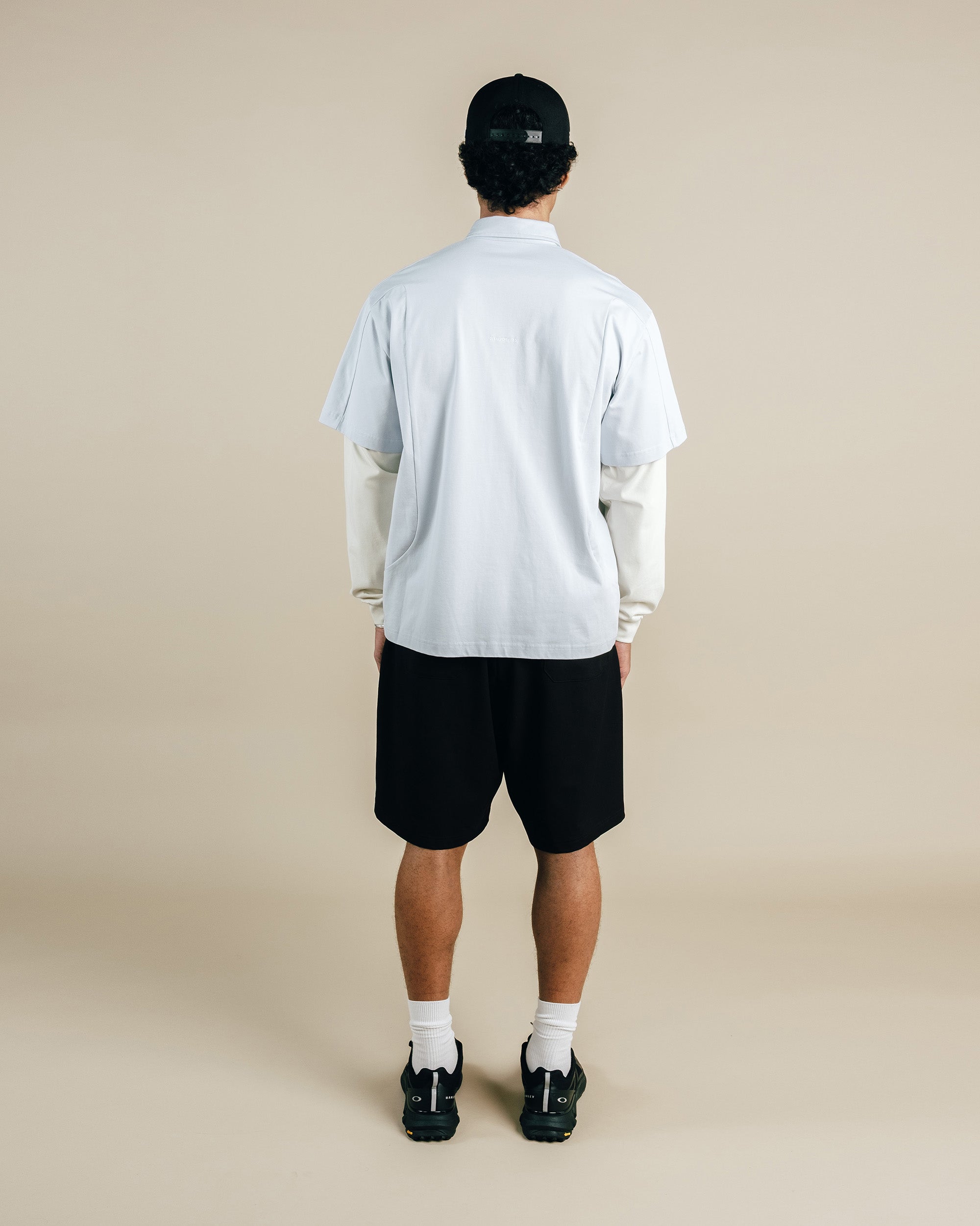 Person wearing a light gray shirt, black shorts, and a black cap on a beige background