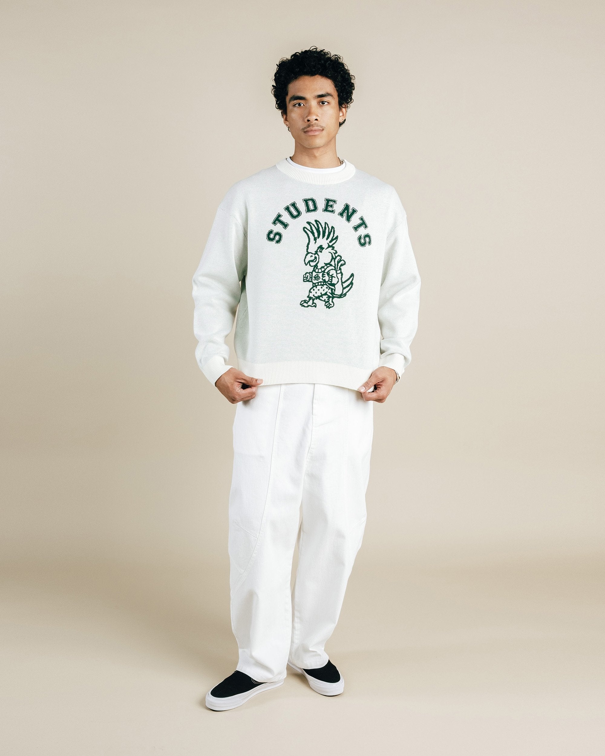 Person wearing a white sweatshirt with 'STUDENTS' text and graphic design, paired with white pants on a beige background.