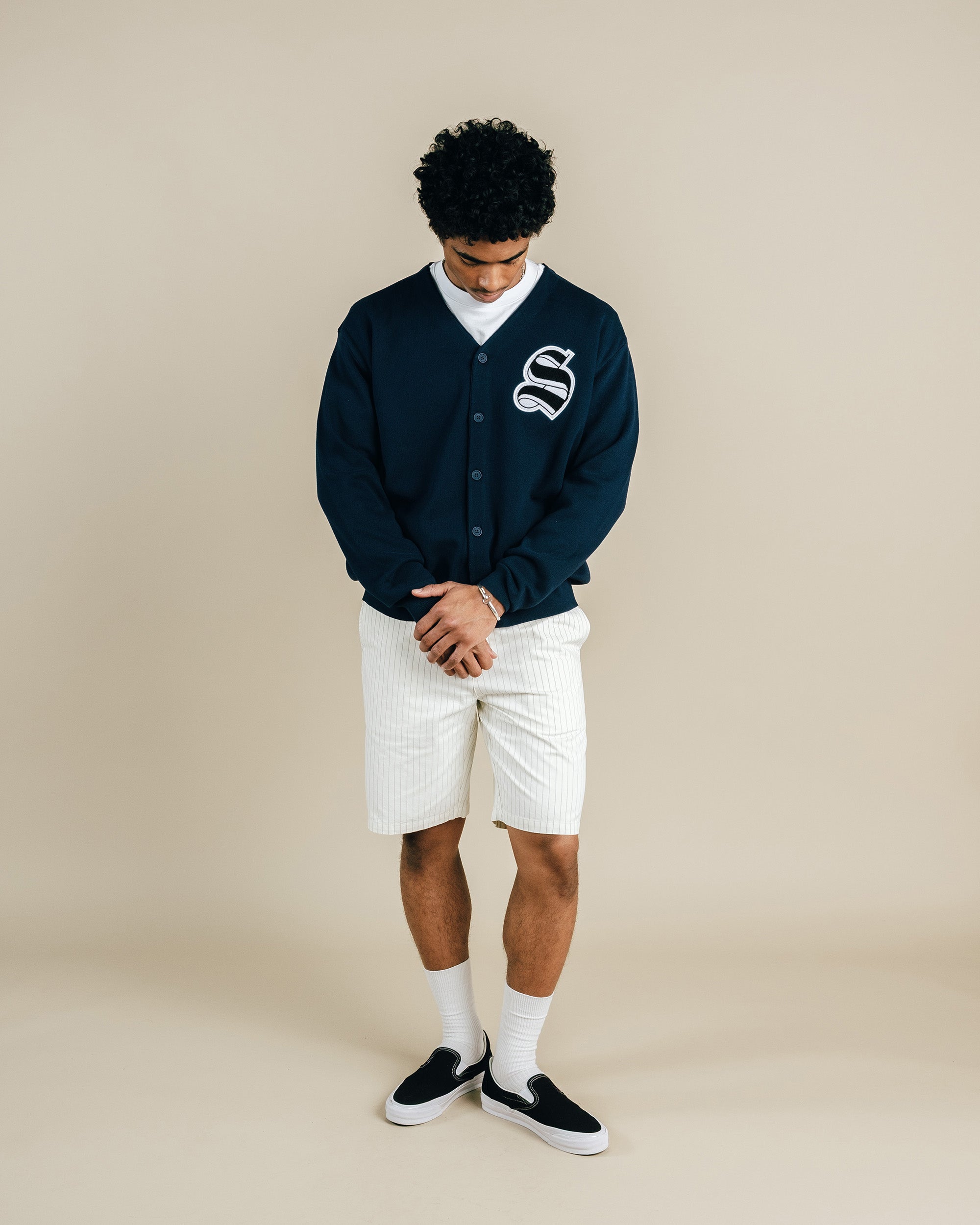 Person wearing a navy cardigan with a logo, white shirt, shorts, and black shoes on a beige background