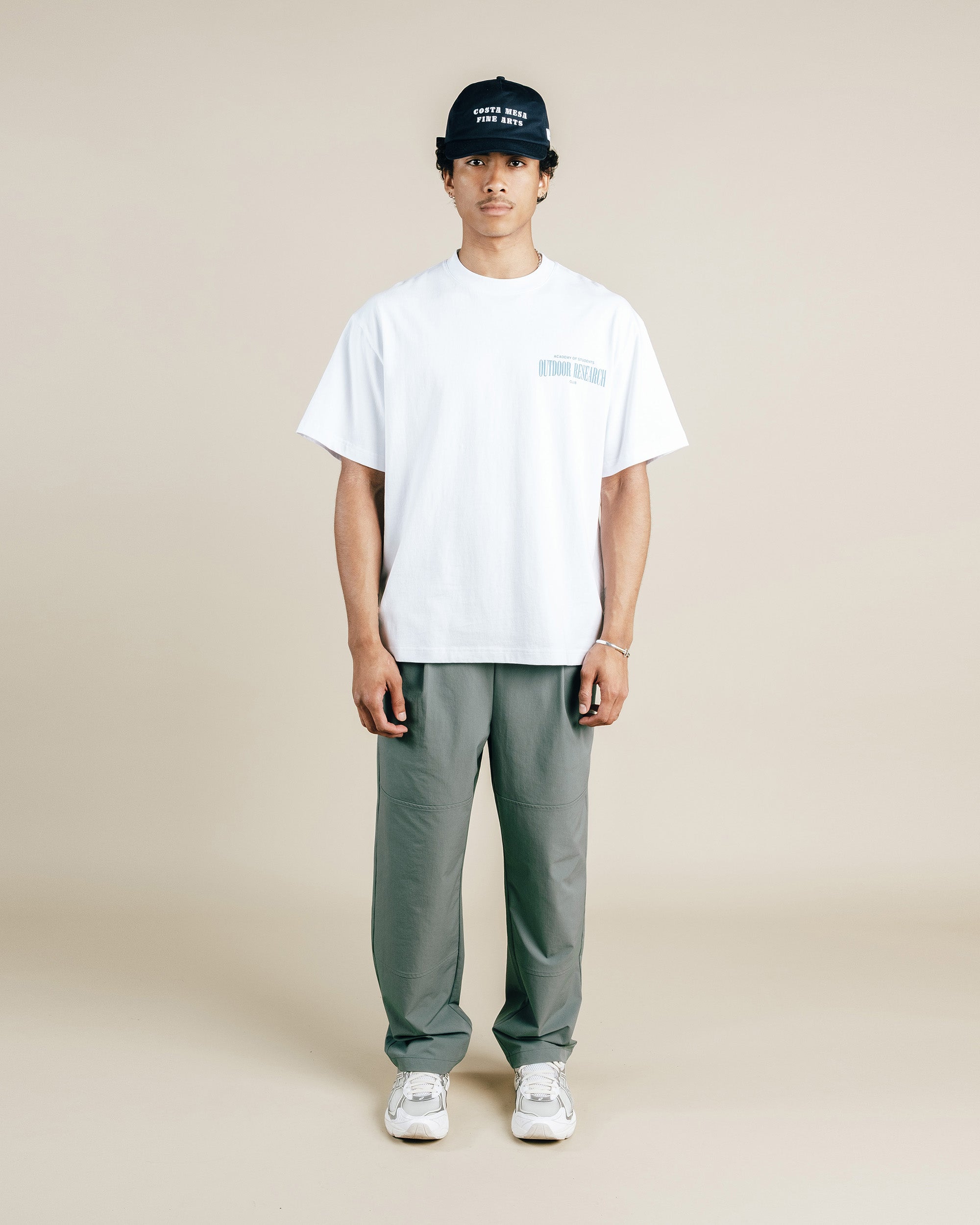 Person wearing a white t-shirt, gray pants, and a black cap on a beige background