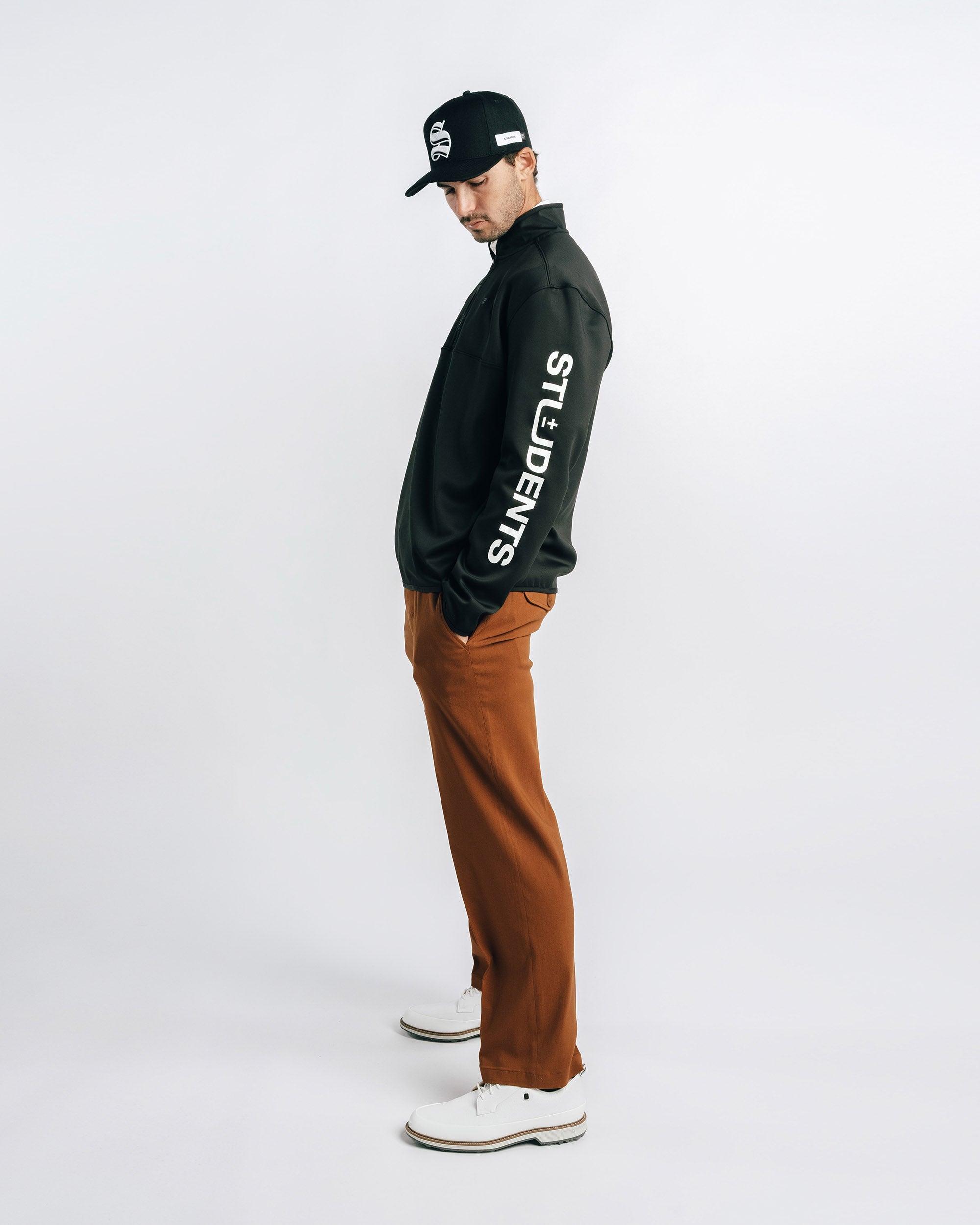 Person wearing a black jacket with white text, brown pants, and a black cap on a white background