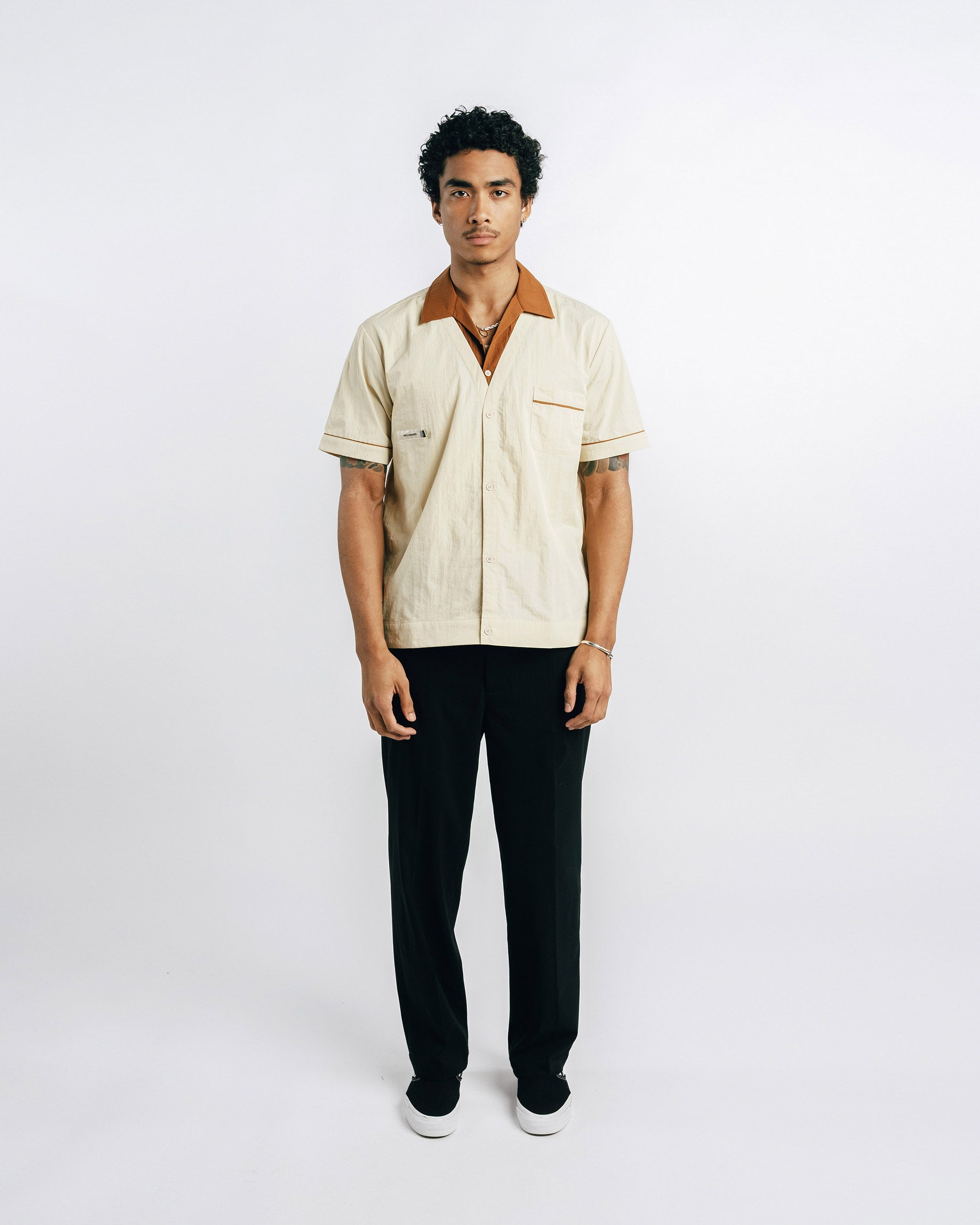 Man wearing a beige shirt with a brown collar and black pants on a white background