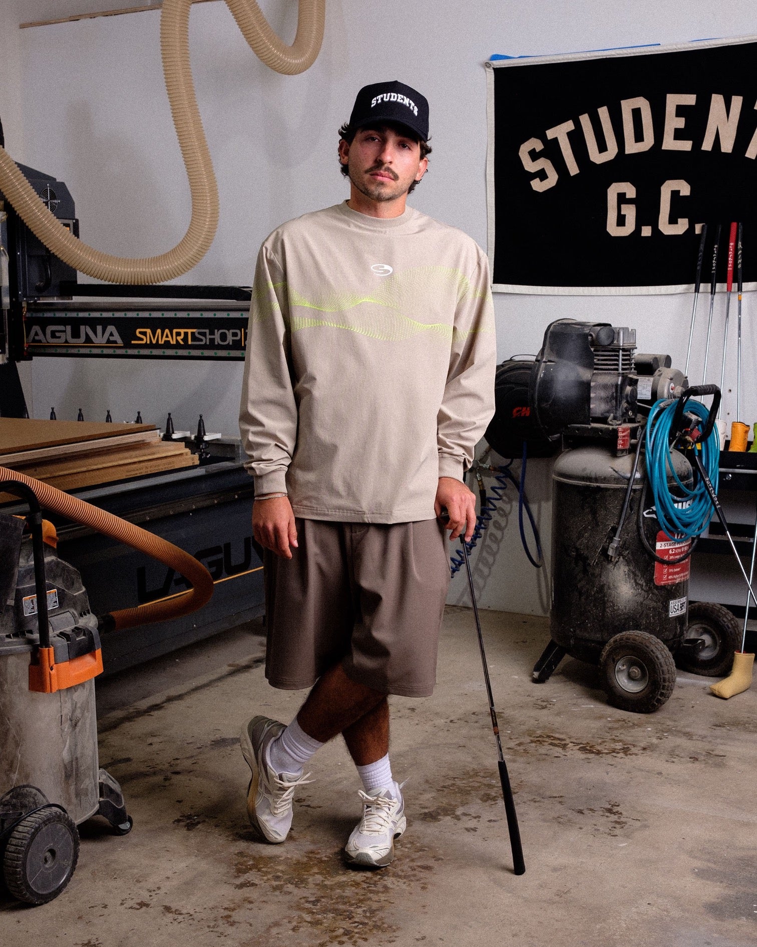 Man standing in a workshop wearing a beige long-sleeve shirt and gray shorts.