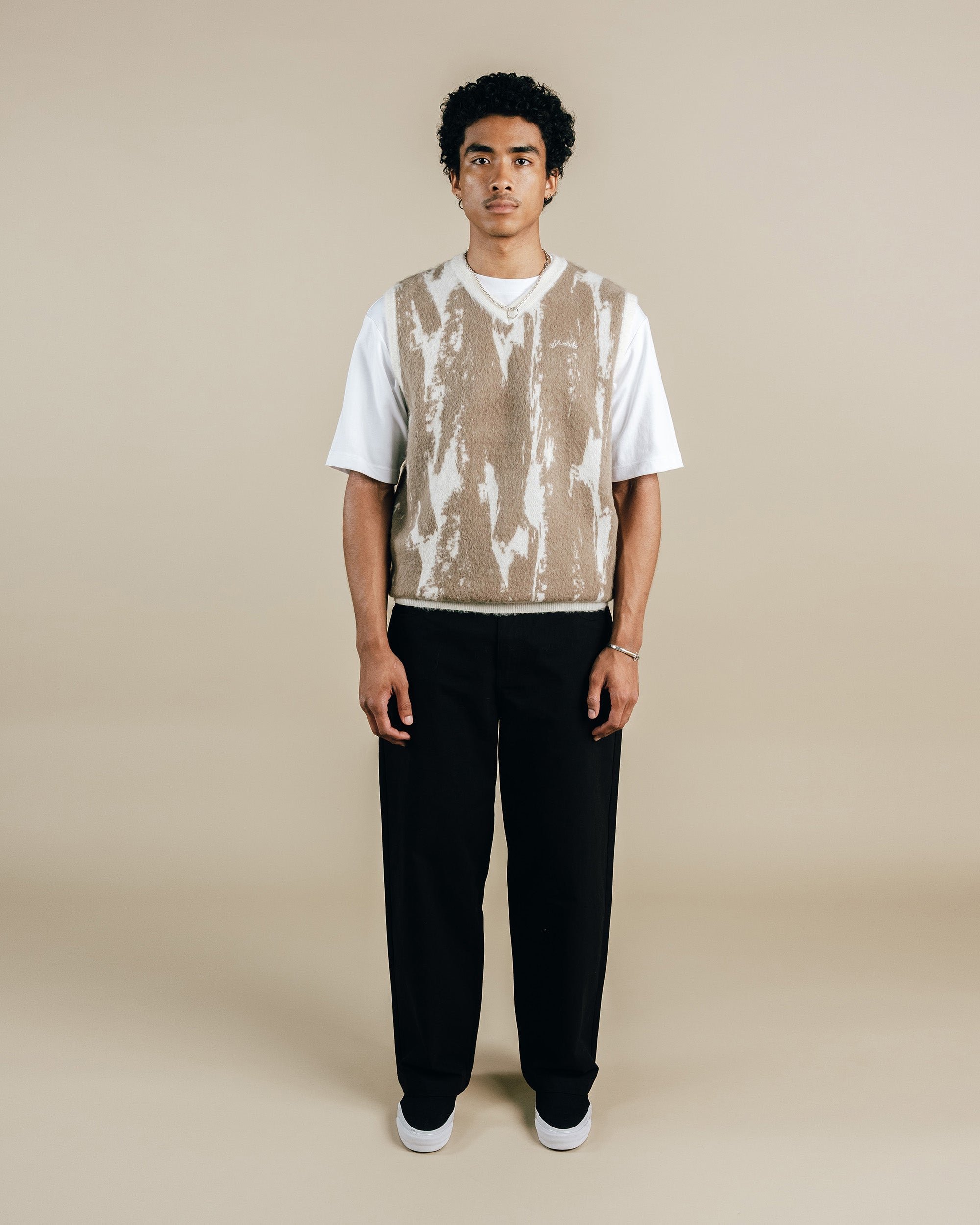 Man wearing a white t-shirt with a patterned vest over it, black pants, and white sneakers on a beige background.