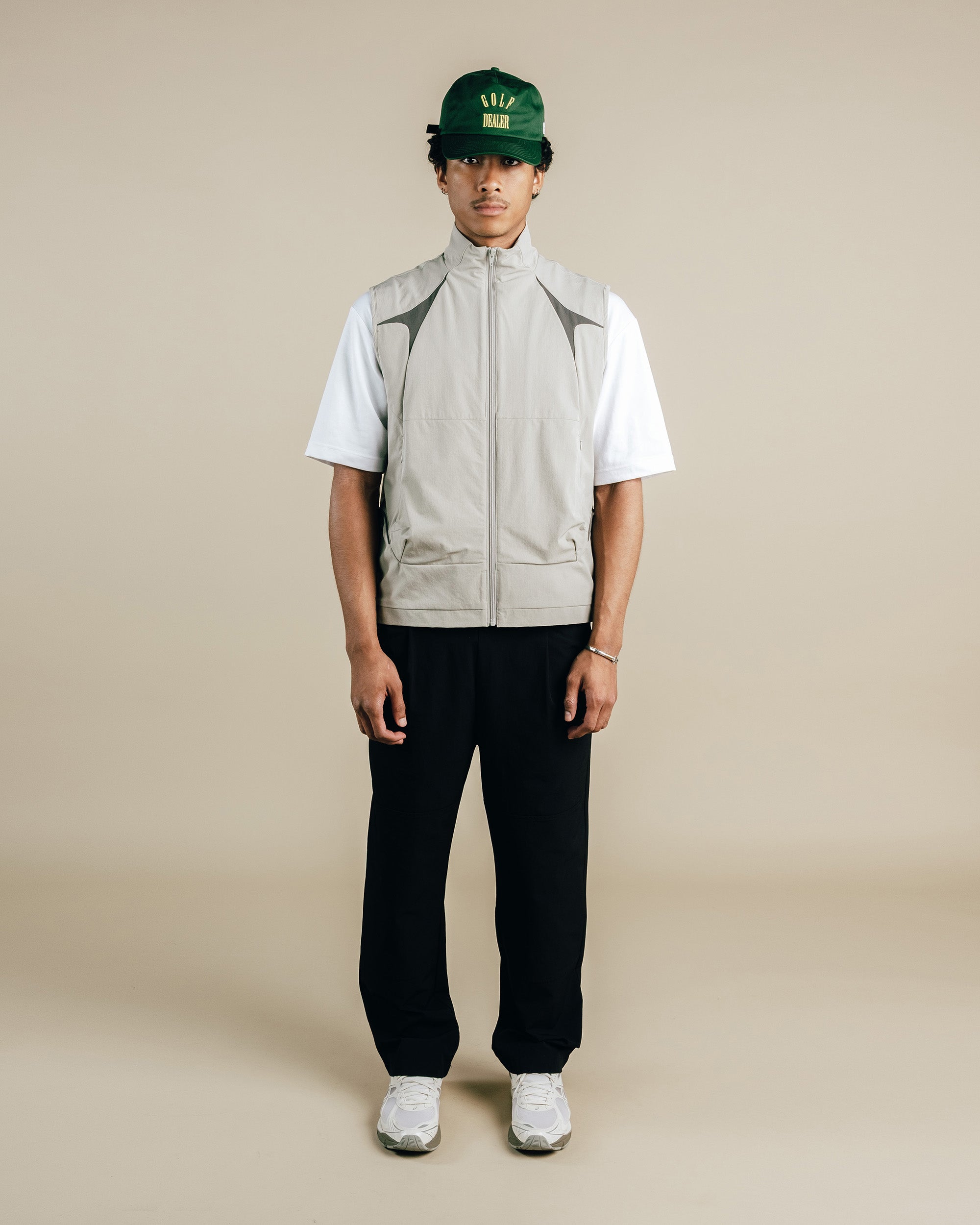 Person wearing a beige vest, white shirt, black pants, and green cap on a beige background