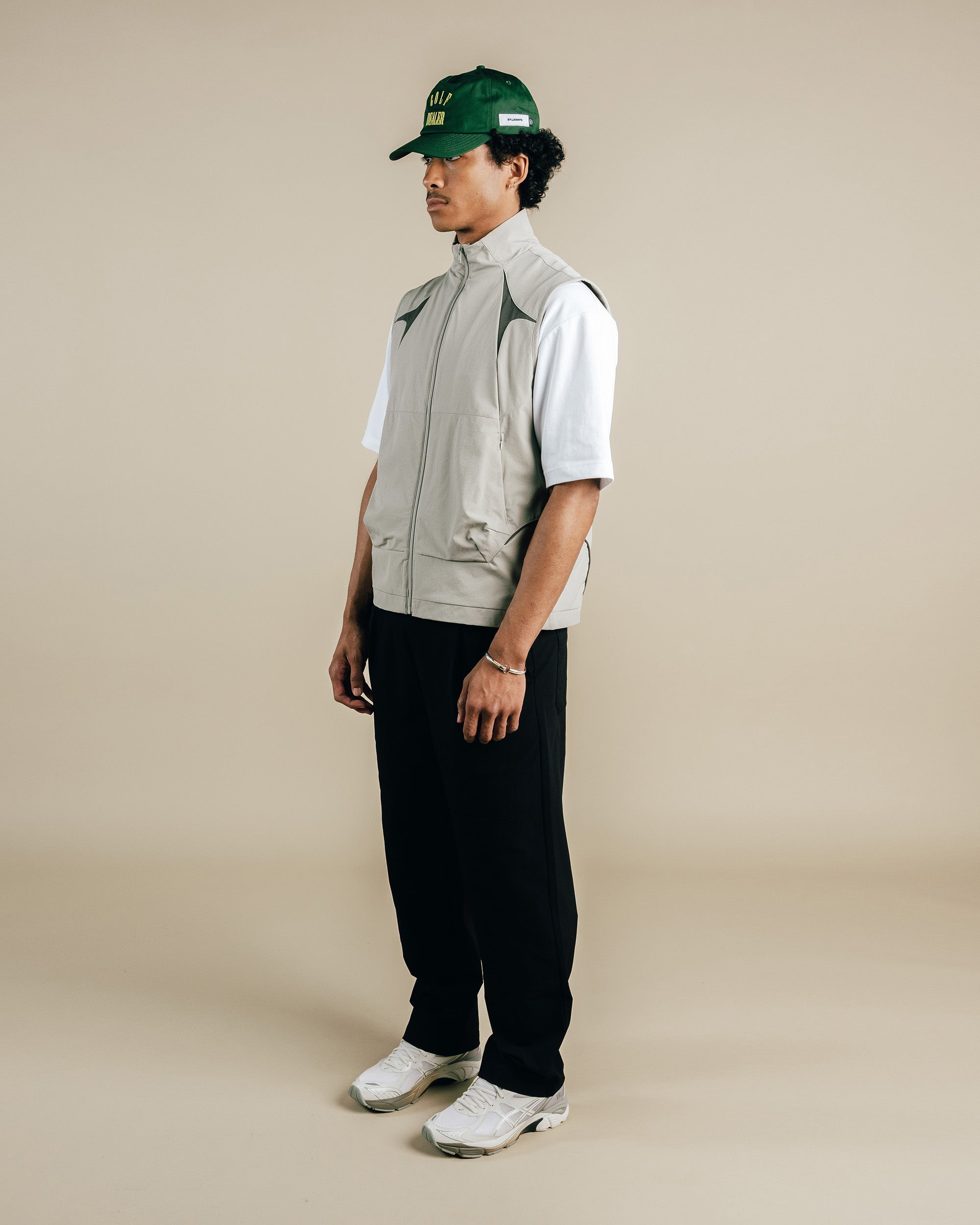 Person wearing a green cap, light gray vest, white shirt, black pants, and white sneakers on a beige background