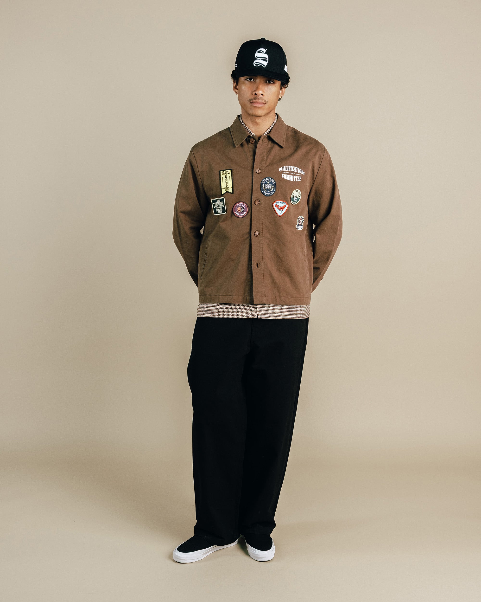 Person wearing a brown jacket with patches and black pants on a beige background