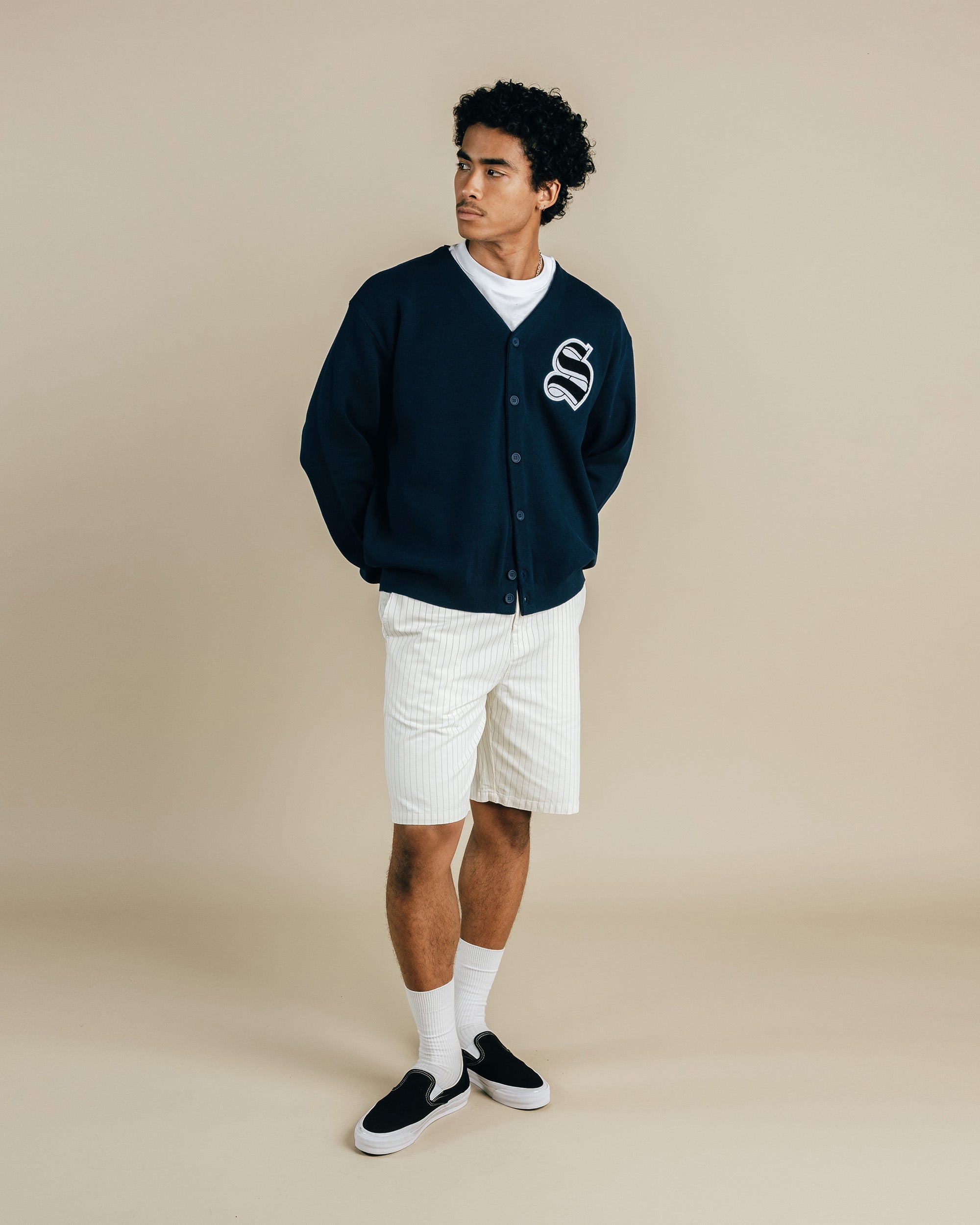 Man wearing a navy cardigan with a logo, white shirt, shorts, and black shoes on a beige background