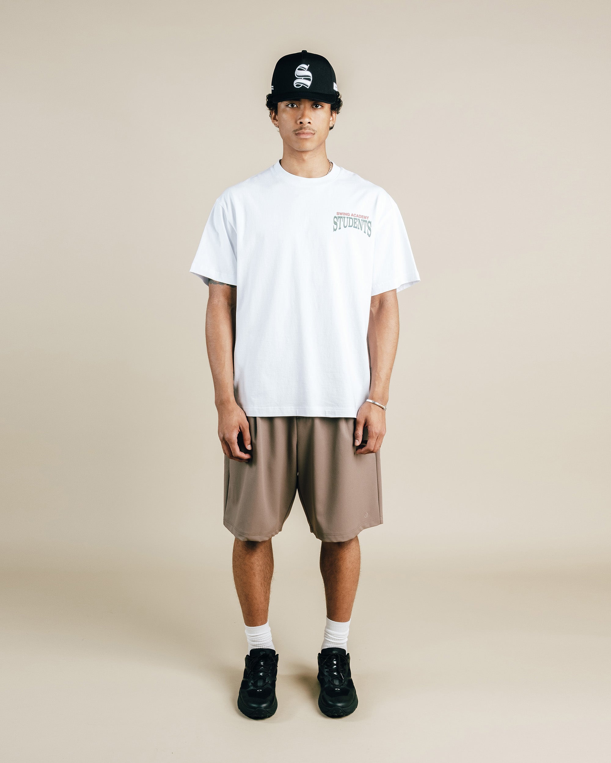 Person wearing a white t-shirt, beige shorts, black sneakers, and a black cap on a beige background