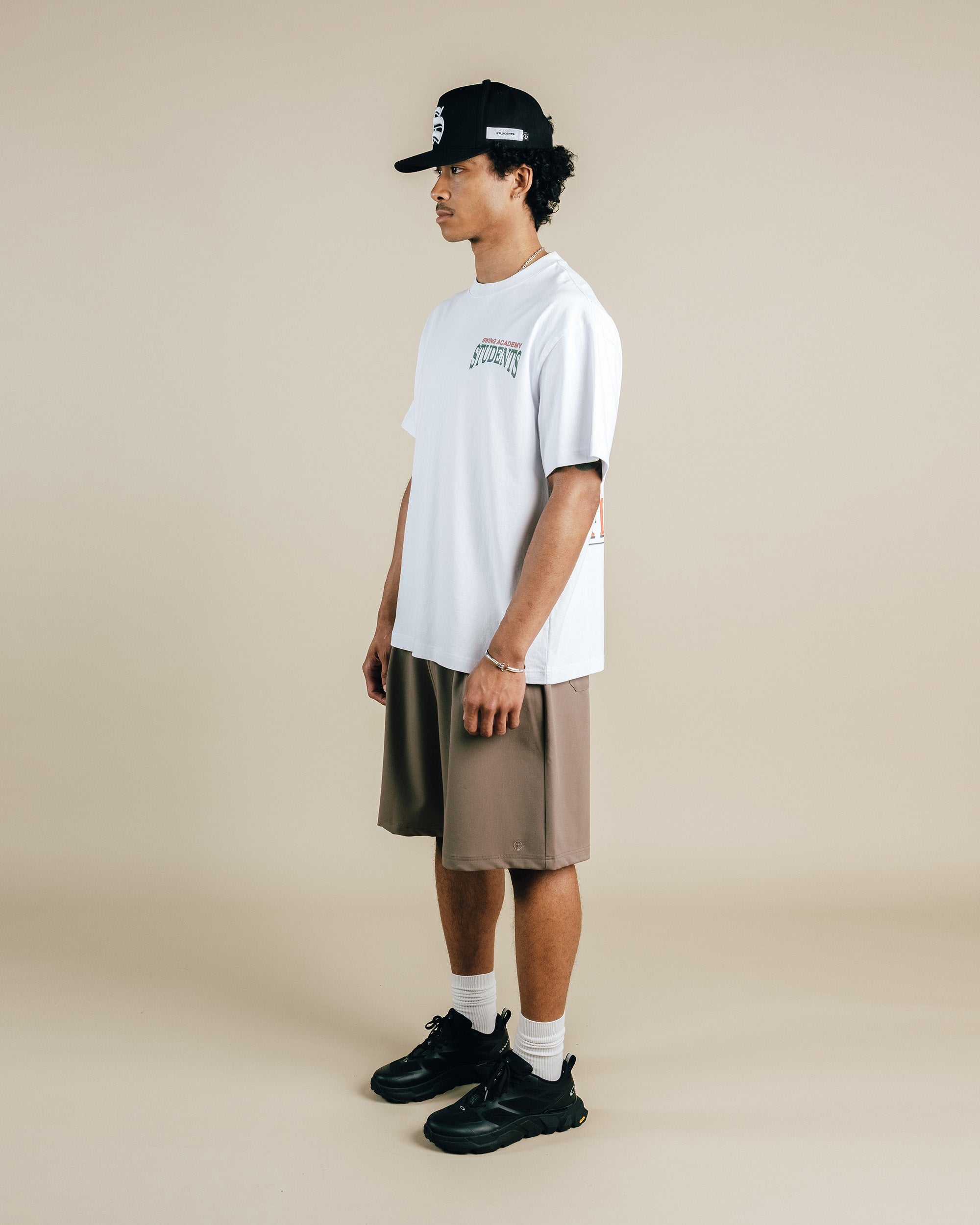 Person wearing a white t-shirt, beige shorts, black sneakers, and a black cap on a beige background