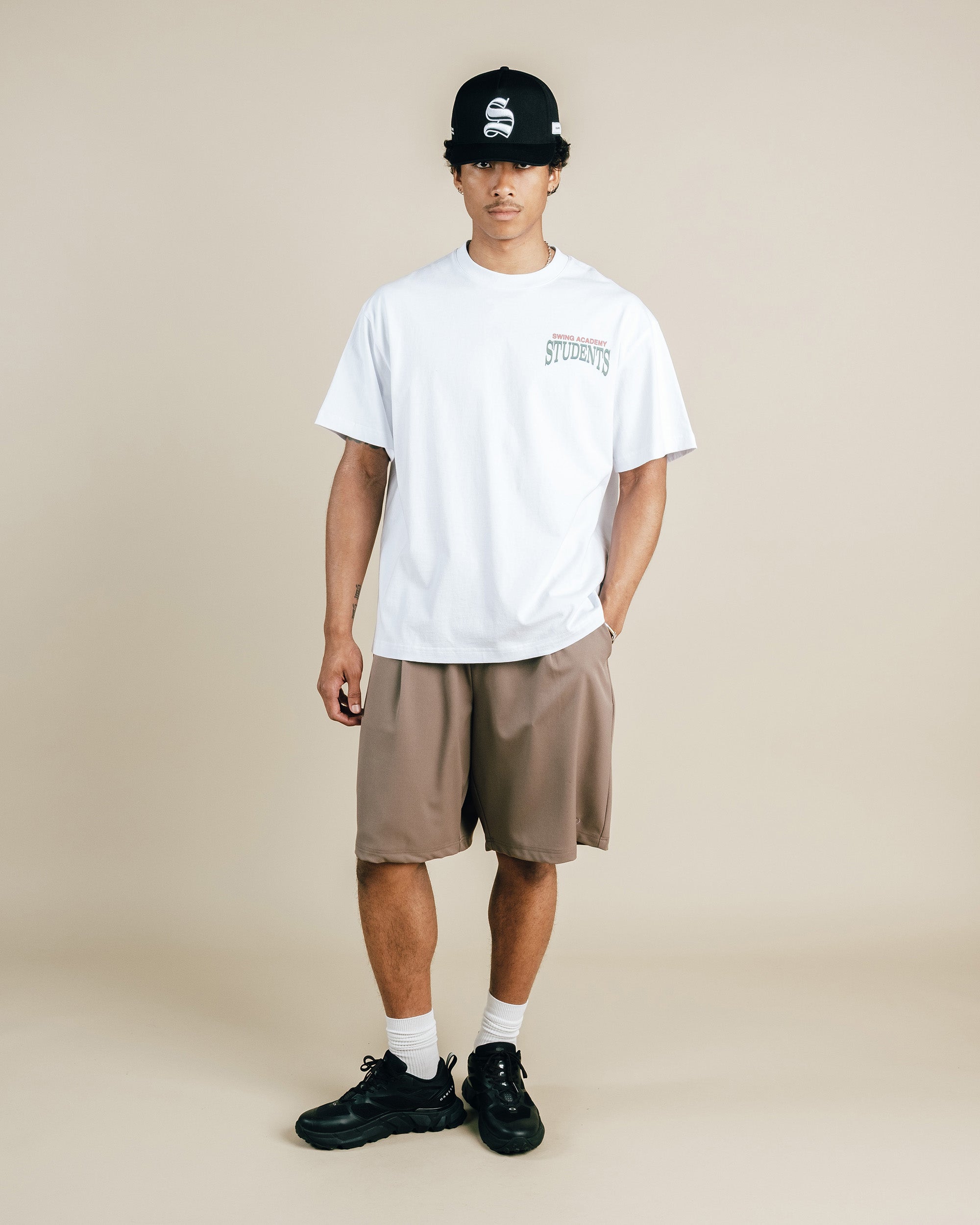Person wearing a white t-shirt, beige shorts, black shoes, and a black cap on a beige background