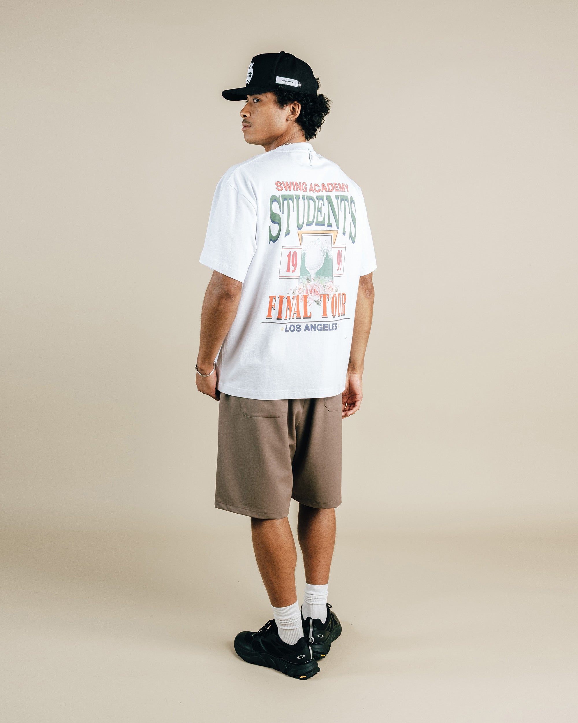 Person wearing a white t-shirt with graphics and text, brown shorts, and black cap on a beige background