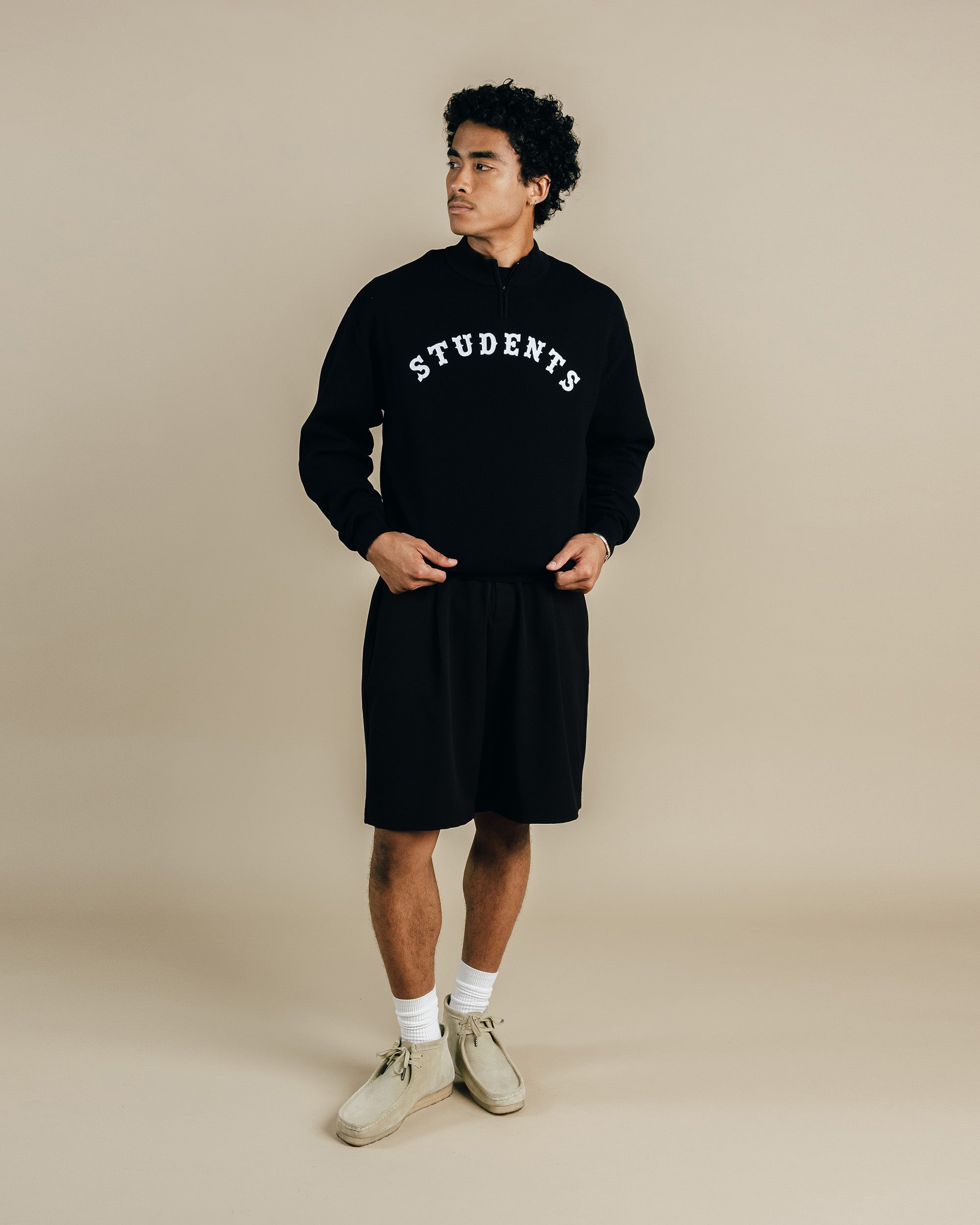 Person wearing a black hoodie with 'STUDENTS' text and black shorts on a beige background