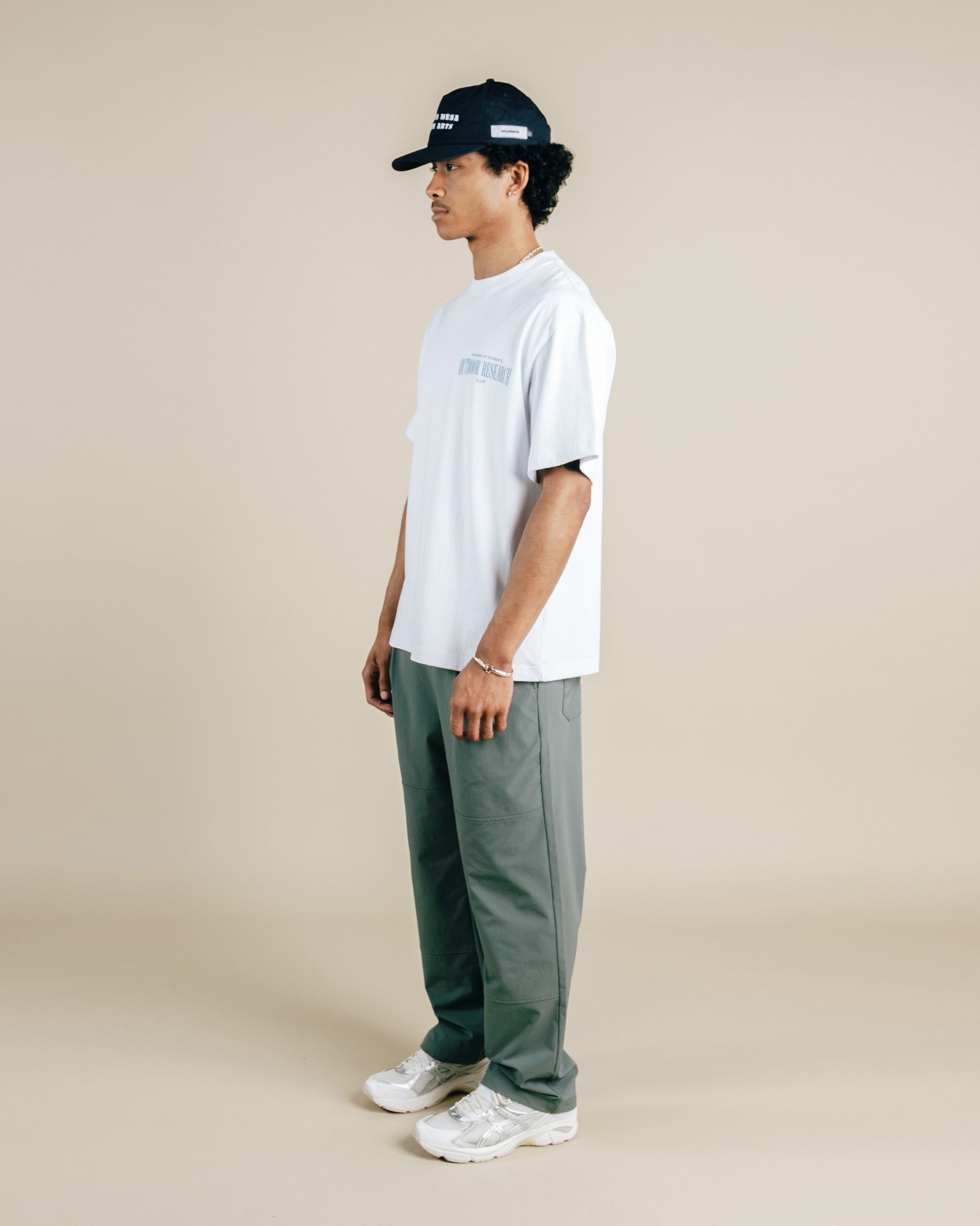 Person wearing a white t-shirt, green pants, and a black cap on a beige background