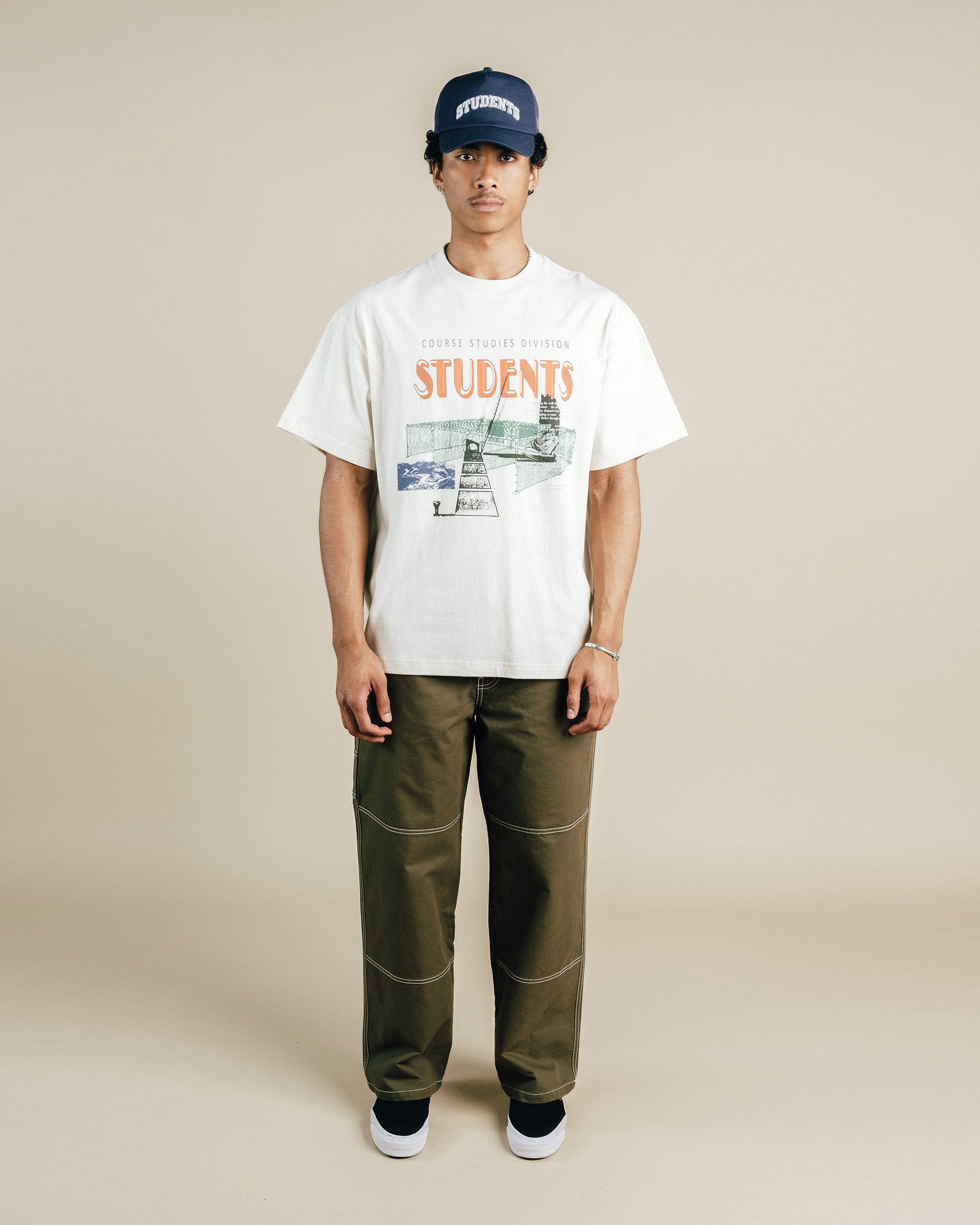 Person wearing a nude t-shirt with graphics and text, green pants, and a navy cap on a beige background